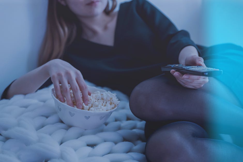 A person sitting on a couch in front of a television late at night, with their eyes glued to the screen and a bowl of popcorn on their lap.